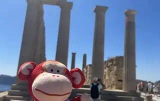Colorful plush monkey toy with "Monkey Puzzle Day Nurseries" logo, against ancient Greek ruins, sunny sky, and visitors, highlighting childcare, early education, and nursery services.