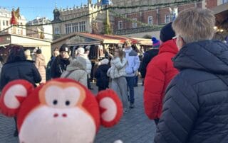 Colorful monkey plush toy at Christmas market outdoor scene during winter season with festive decorations, crowded visitors, and historic architecture in the background.