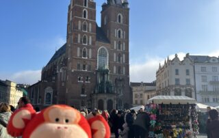 Colorful monkey plush toy in front of St. Mary's Basilica in Krakow, Poland, during a busy market day, showcasing a fun and vibrant atmosphere perfect for children's toys and educational activities at Monkey Puzzle Day Nurseries.