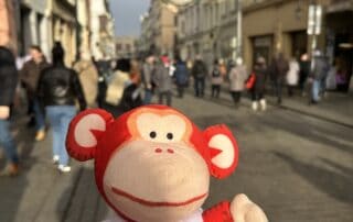 Monkey puzzle day nursery plush toy monkey wearing branded shirt in city street with people walking in background.