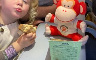 Bright young girl enjoying a snack on an airplane with a plush monkey "Monkey Puzzle" toy and a veggie-themed sandwich, highlighting fun, nutritious meals for children at Monkey Puzzle Day Nurseries.