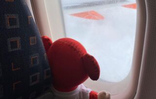 Fluffy red teddy bear looking out of an airplane window at the snowy landscape and runway during winter travel.
