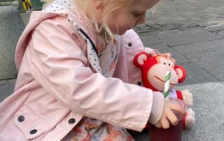 Bright young girl with blonde hair, wearing a pink jacket and floral dress, sitting on a park bench at Monkey Puzzle Day Nurseries, playing with a plush monkey toy and a drink.