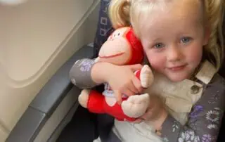 Soft toy monkey hugging young girl on airplane seat, cheerful child with blond hair in pigtails, engaging in travel adventure at Monkey Puzzle Day Nurseries.