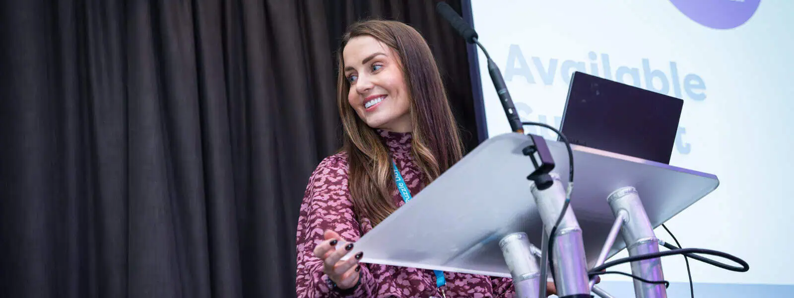 Bright young woman presenting at a professional event, standing behind a podium with a laptop, on a stage with a dark curtain background, demonstrating confidence in early childhood education.