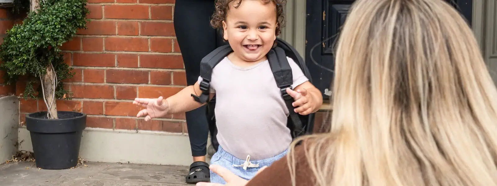 Happy young child with a backpack smiling at a caregiver outside Monkey Puzzle Day Nurseries, emphasizing early childhood education and welcoming environment.