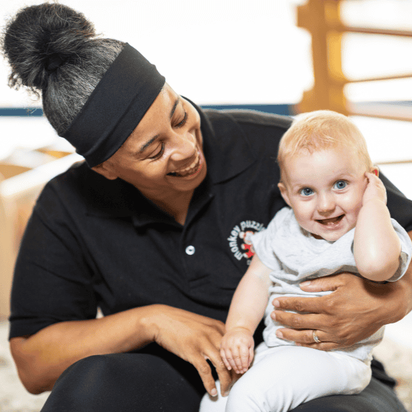 Smiling young child with blonde hair and blue eyes sitting on caregiver’s lap, both smiling warmly in a bright, welcoming nursery environment.