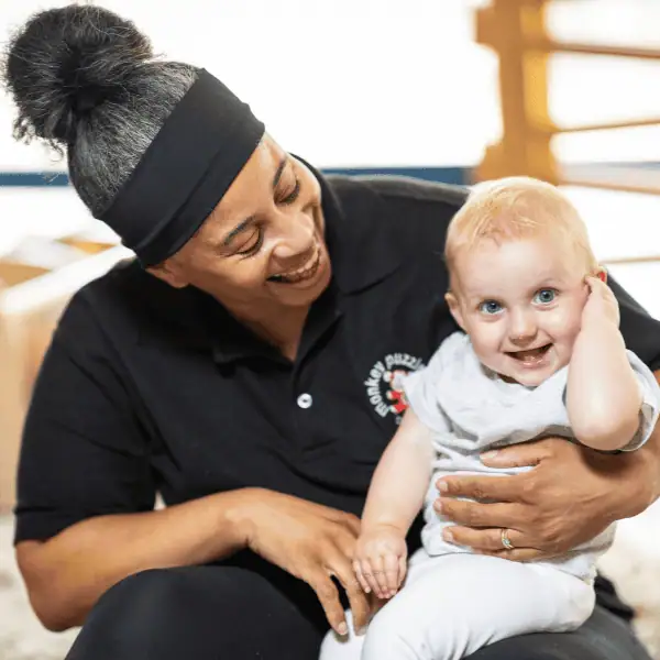 Smiling young child with blonde hair and blue eyes sitting on caregiver’s lap, both smiling warmly in a bright, welcoming nursery environment.