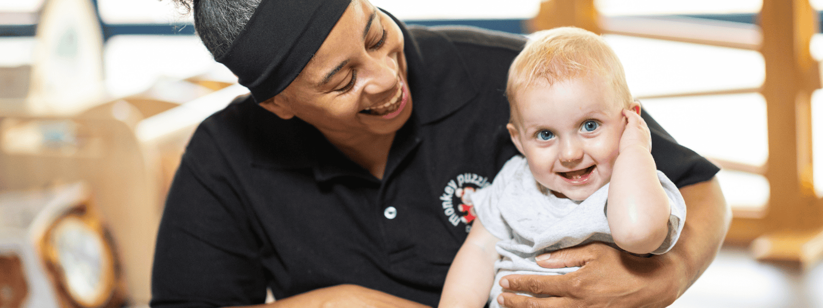 A joyful young child with blonde hair and blue eyes smiling while being held by a caregiver at Monkey Puzzle Day Nurseries, emphasizing a safe and nurturing environment for children.