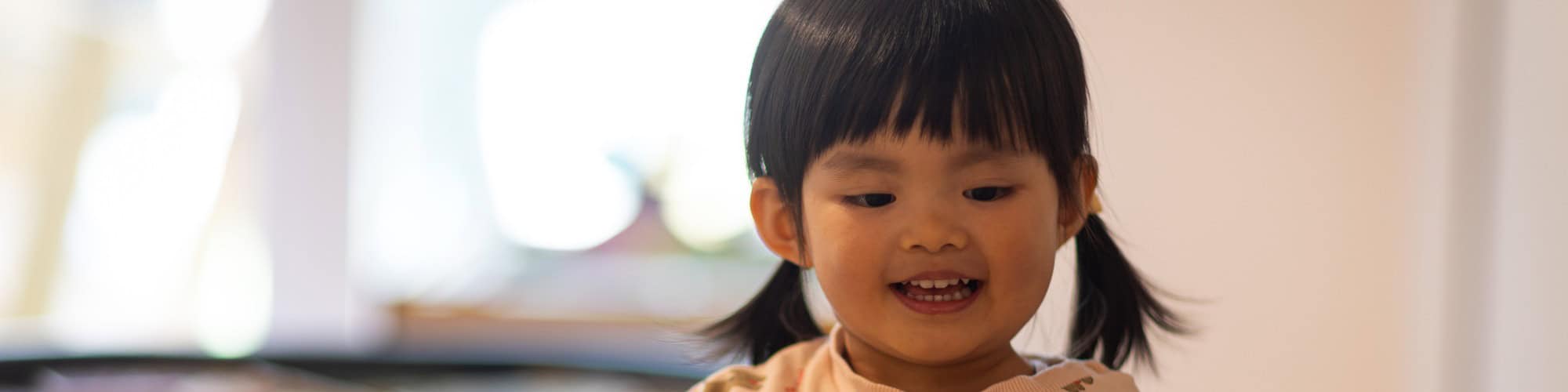 Smiling young girl with pigtails attentively listening in a bright nursery setting, promoting early childhood development and communication skills.