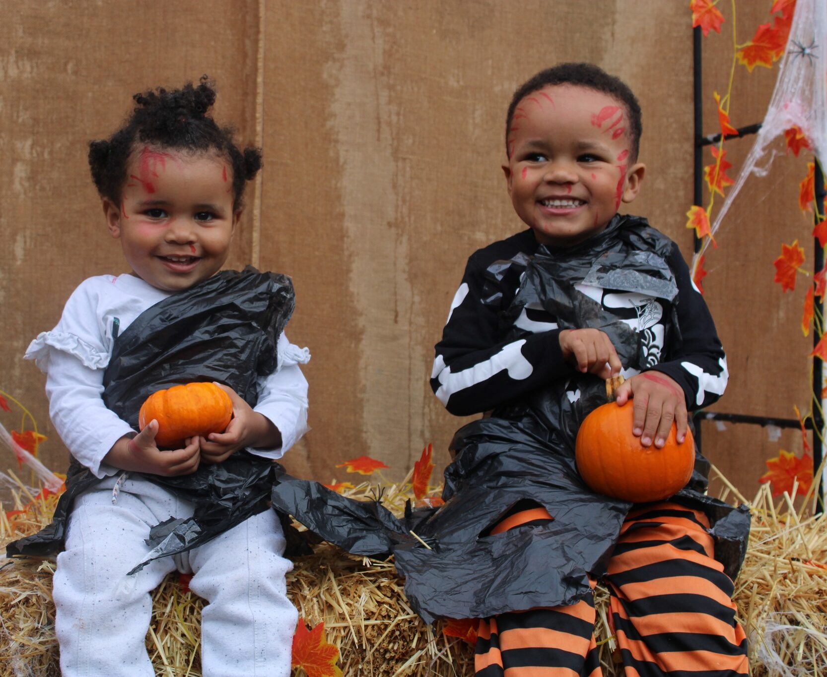 Two children in Halloween costumes sitting on hay bales, holding pumpkins, with autumn decorations in the background.