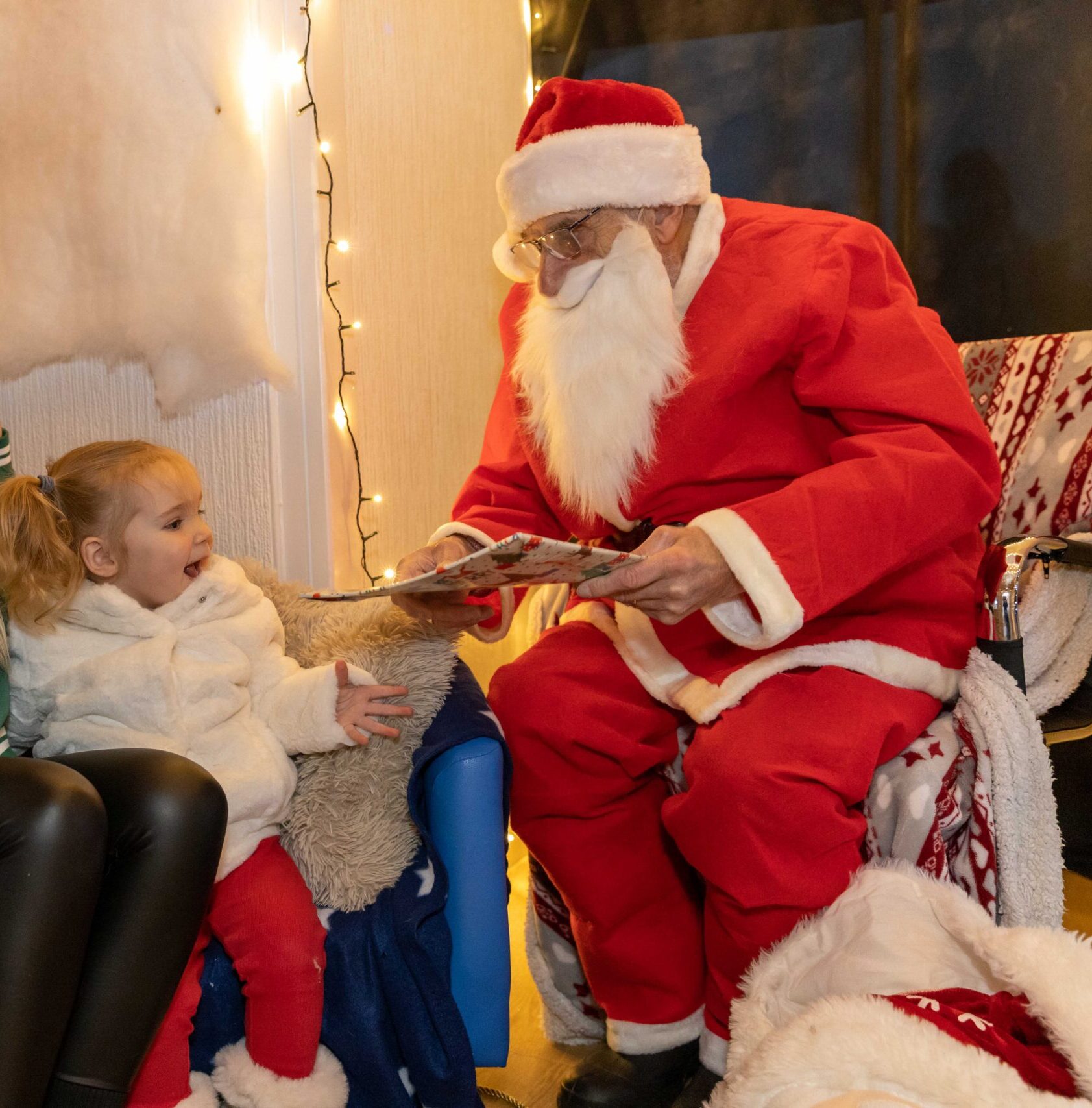 Santa Claus in red suit and white beard talking to young girl and woman at nursery Christmas event.