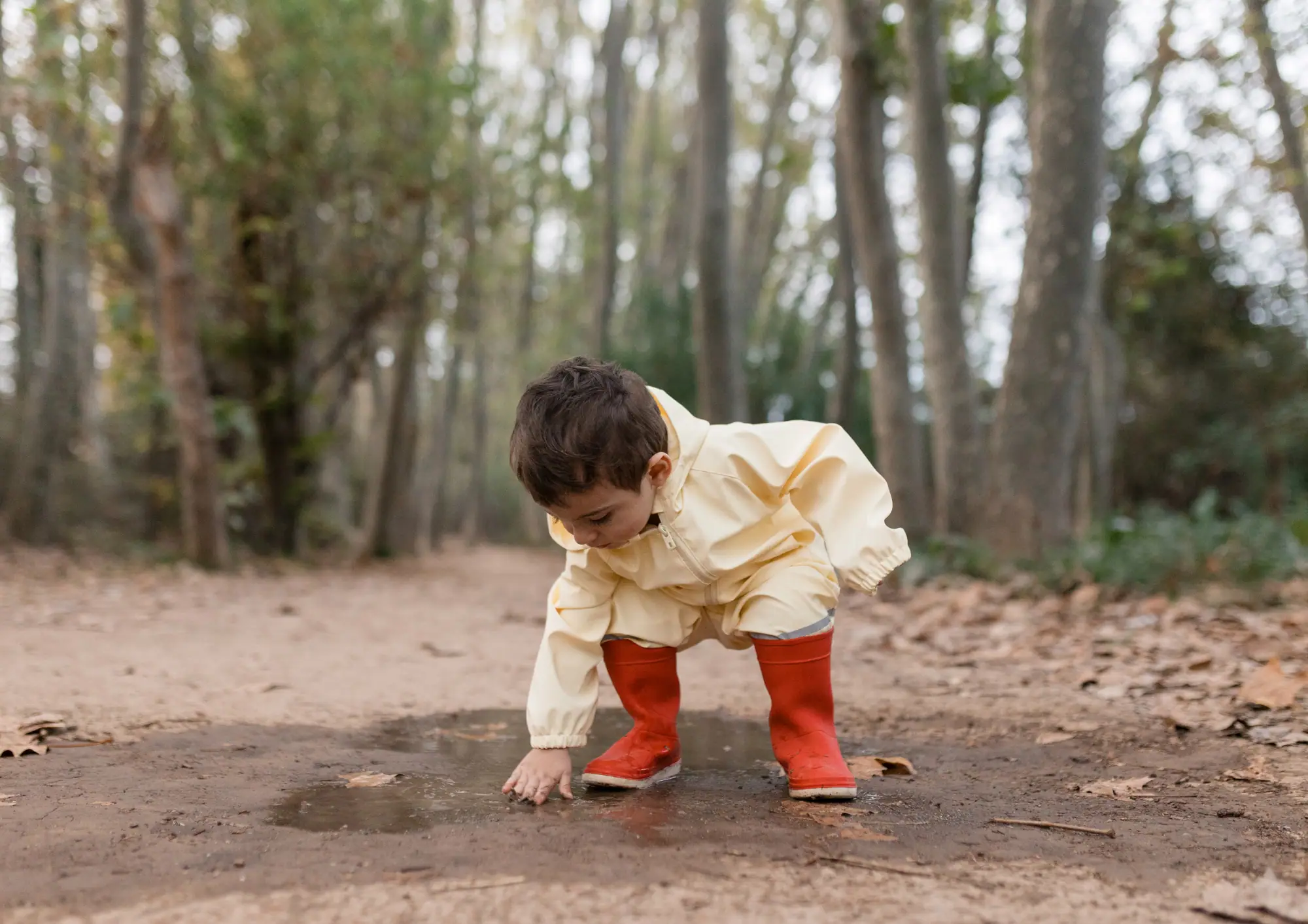 Young child in yellow jacket and red boots exploring muddy ground in a forest setting during winter.